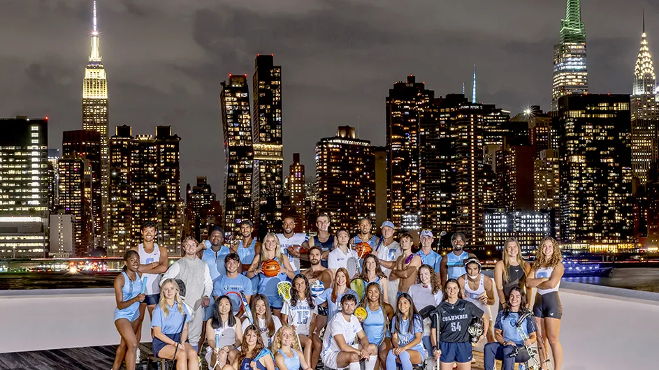 Thirty-one college student athletes in Columbia University sports uniforms pose in front of the illuminated New York City skyline at night. 