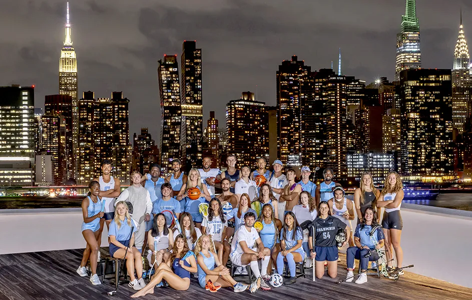 Thirty-one college student athletes in Columbia University sports uniforms pose in front of the illuminated New York City skyline at night. 