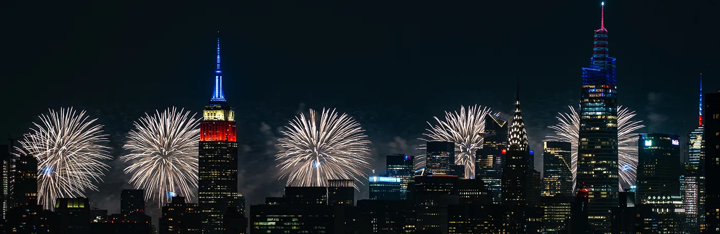 New York City skyline of Empire State building, Chrysler Building, and other skyscrapers with white fireworks illuminating the sky on July 4th. 