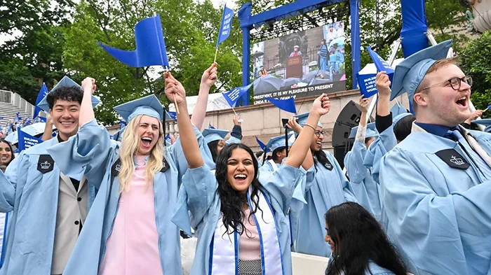 Mailman School of Public Health graduates celebrate with blue flags waving in the air.