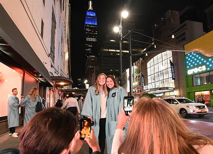 Parents take photos of two friends in Columbia graduation gowns with the Empire State Building in the background