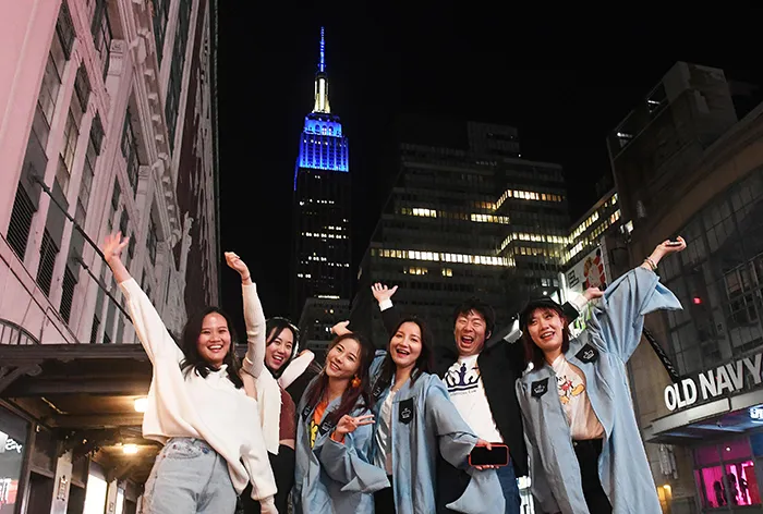 A group of friends in Columbia graduate gowns poses at 34th Street and 7th Avenue with the Empire State Building in the background.
