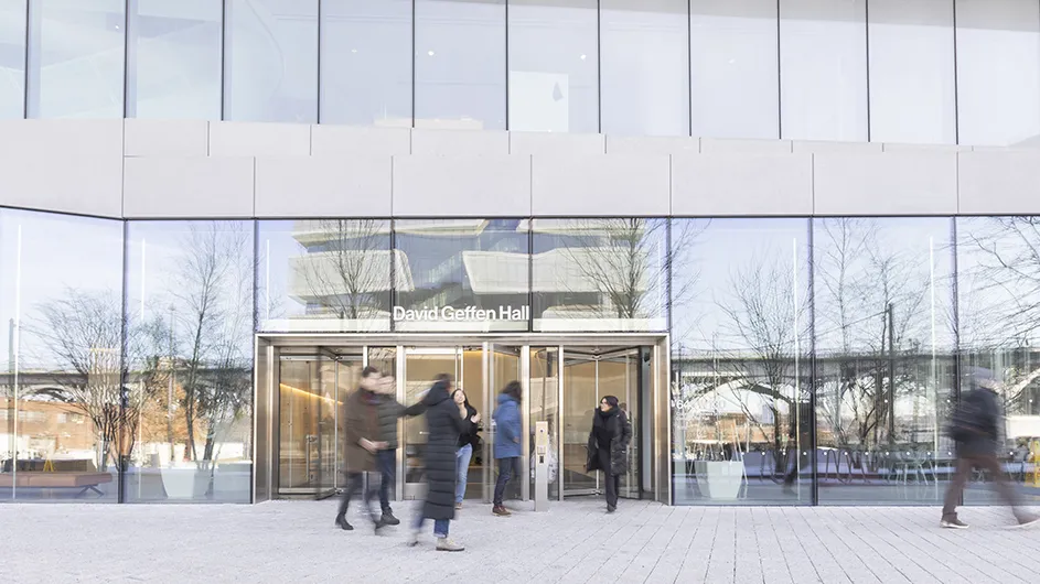 Several university students walk near David Geffen Hall with its façade showing a reflection of Henry R. Kravis Hall across the square at Columbia Business School on a cold sunny day. 