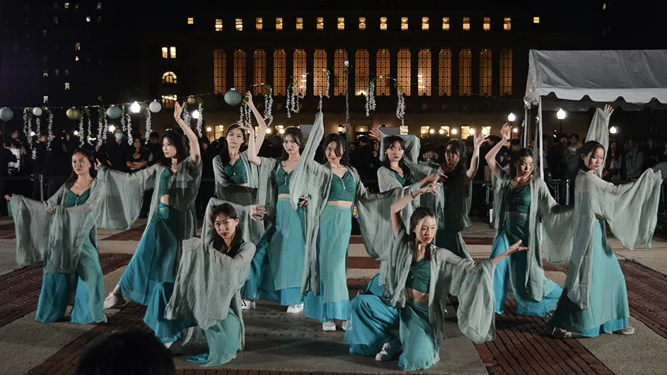 Eleven university students in draped green shawls and long shirts perform a traditional Chinese dance on Low Plaza with spectators and illuminated Butler Library in the background at Columbia University at night. 