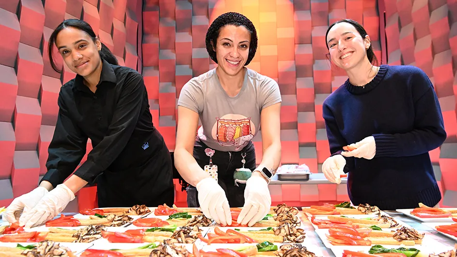 Three Columbia Dining staff members smile and pose for the camera while preparing plates of sushi-prep food at Wang Pavilion in Lerner Hall at Columbia University.