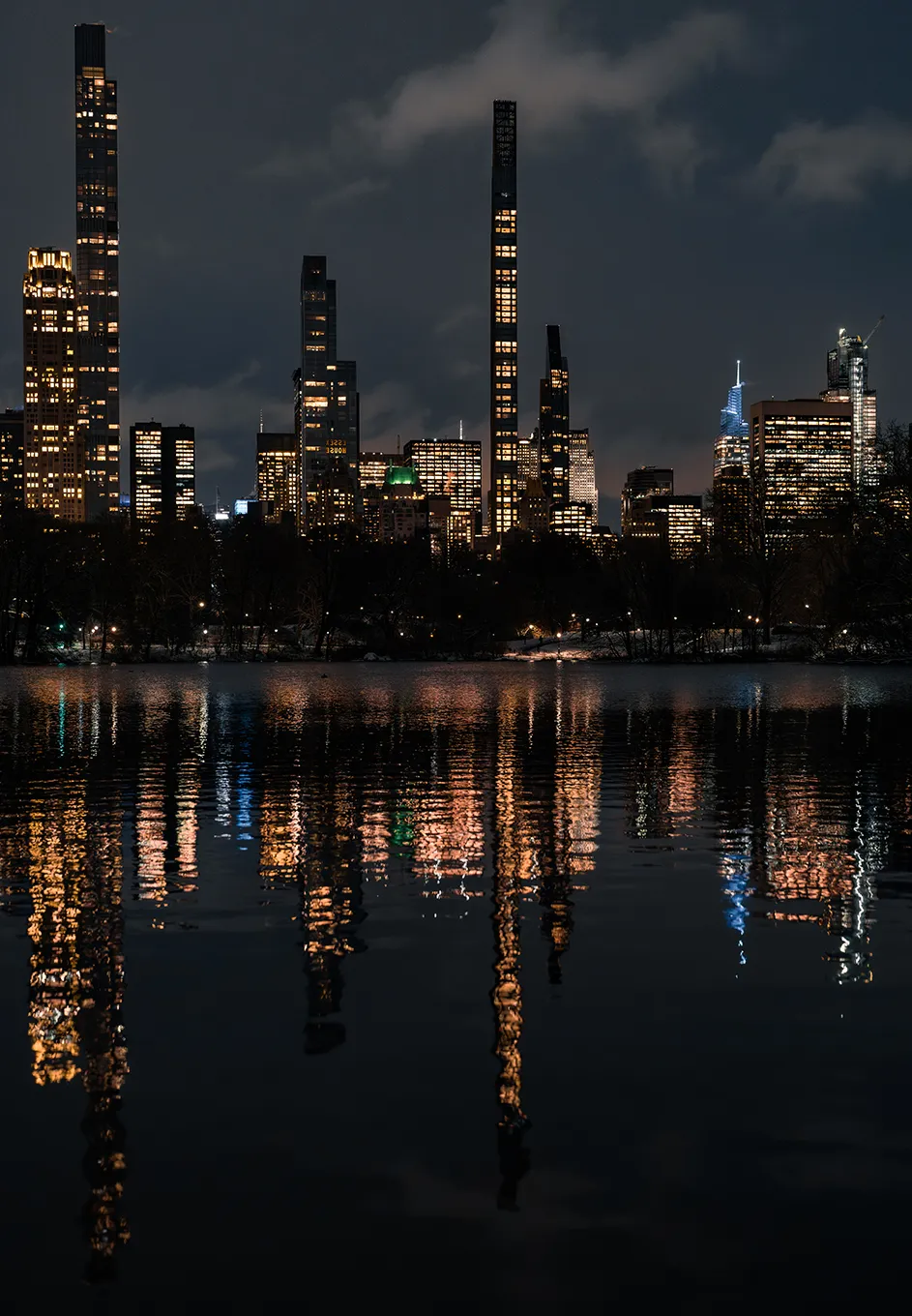 Illuminated midtown Manhattan skyscrapers reflected on Central Park's pond lined by snowy tree-lined paths on a winter night. 