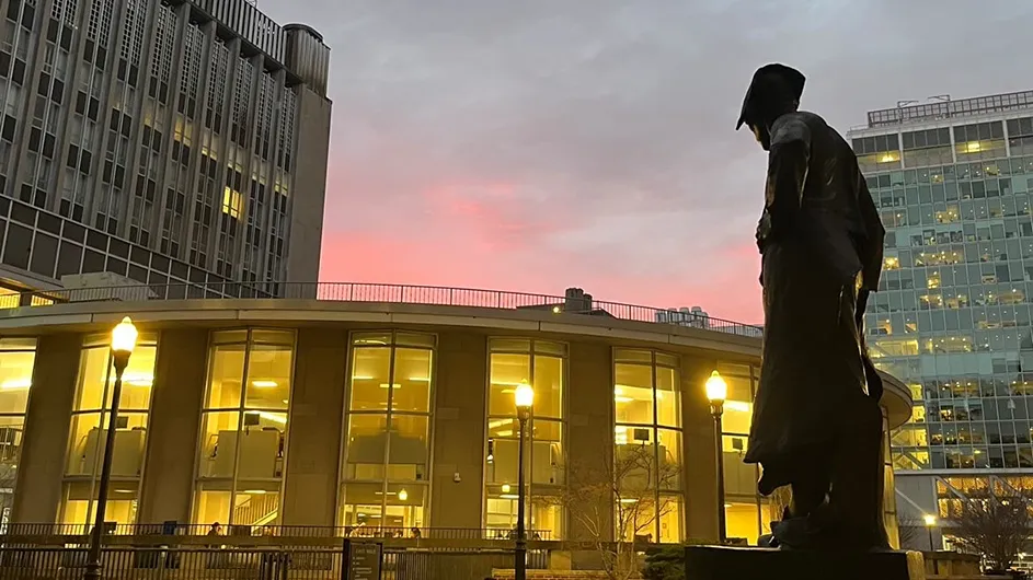 Le Marteleur Hammersmith sculpture looks toward illuminated Uris Hall Library against a pink sunset sky at Columbia University. 