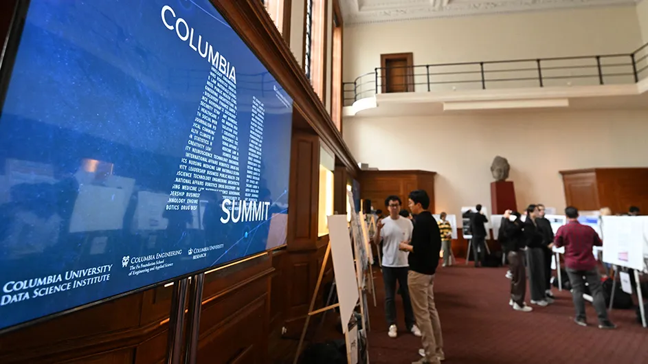 Several university students and researchers discuss poster content near blue branding which reads Columbia AI Summit on a big screen at Columbia University.