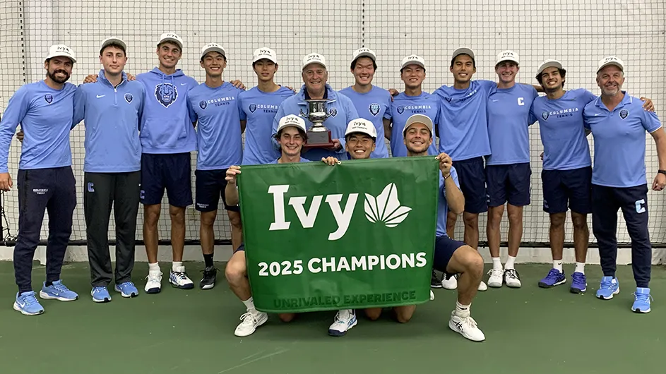 Fifteen men's tennis university players and coaches in Columbia blue uniforms smile and pose with an Ivy 2025 Champions sign at an indoor tennis court.