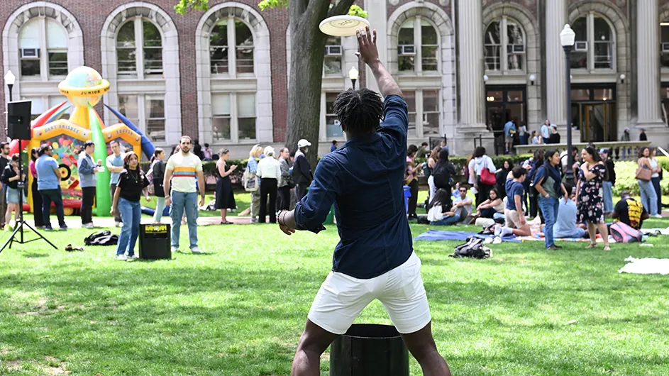 Three university students play a game of frisbee as one student catches a white frisbee on Lewisohn Quad with dozens of other students and staff enjoying a campus block party in the background at Columbia University on a sunny spring day. 