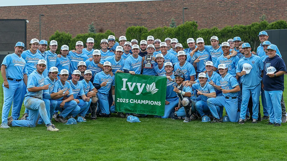 Several men's baseball players and coaches in Columbia blue uniforms smile and pose with a trophy and Ivy League 2025 Champions sign at Columbia's Baker Athletics Complex.