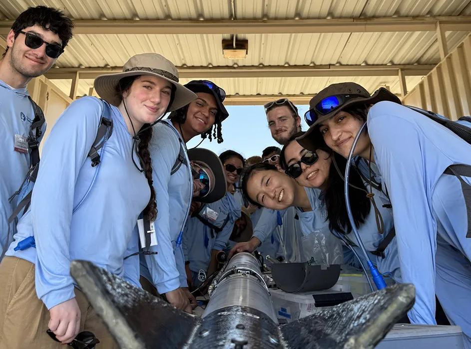 Several Columbia University students in light blue shirts smile and pose with their hybrid rocket in a shed in the Mojave Desert. 