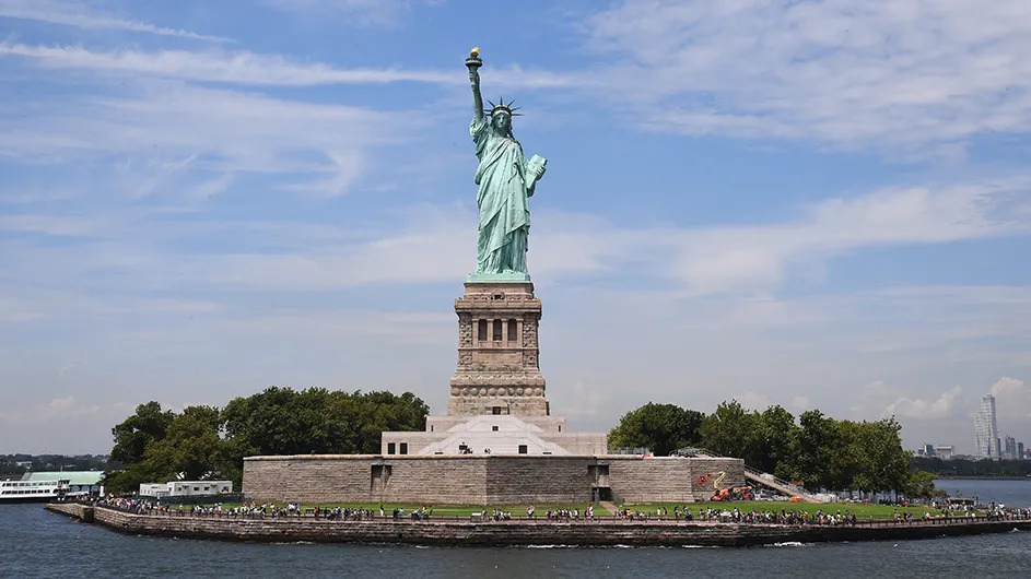 The tall copper Statue of Liberty towers over spectators on Liberty Island in the New York Bay on a sunny day. 