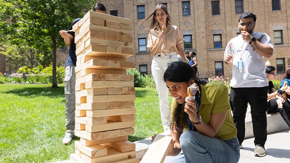 A university staff member holding an ice cream cone in one hand and a jenga block in the other hand considers what the next jenga game move will be as three ice-cream-eating colleagues look on at Columbia University Irving Medical Center's Ice Cream Social on a hot summer day.