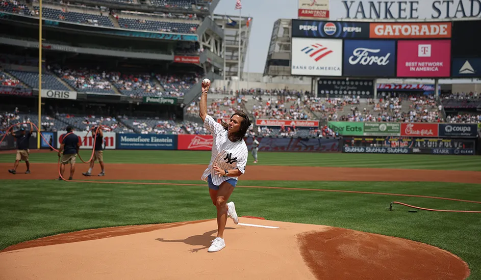 Coach Megan Griffith wearing a New York Yankees jersey throws a baseball pitch from the mound at Yankees stadium on a sunny day. 