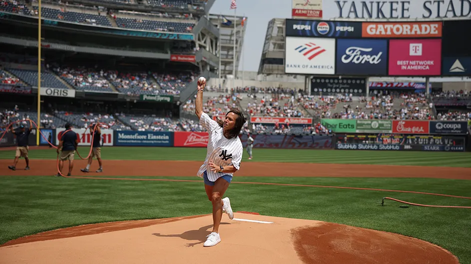 Coach Megan Griffith wearing a New York Yankees jersey throws a baseball pitch from the mound at Yankees stadium on a sunny day. 