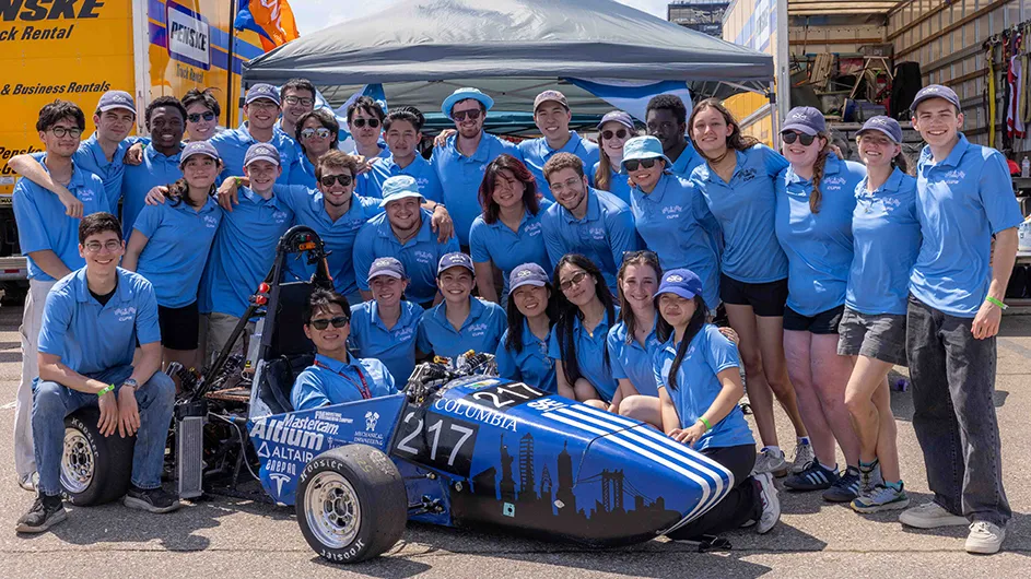 Several Columbia University students in light blue shirts pose and smile with their formula race car at Michigan International Speedway's 2025 Formula Electric Vehicle Competition. 