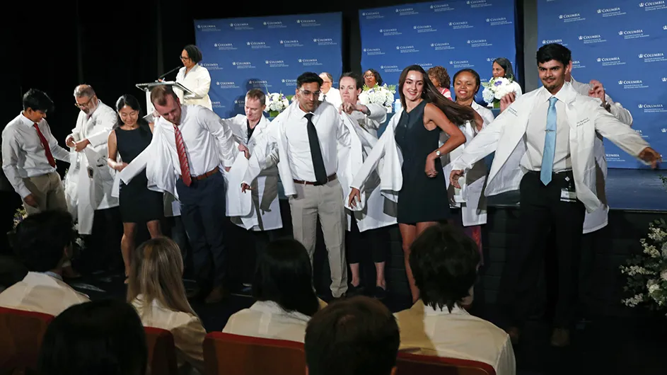Six Columbia University medical students are putting on their white medical coats in front of six faculty members in white coats at Columbia's Vagelos College of Physicians and Surgeons' White Coat Ceremony.