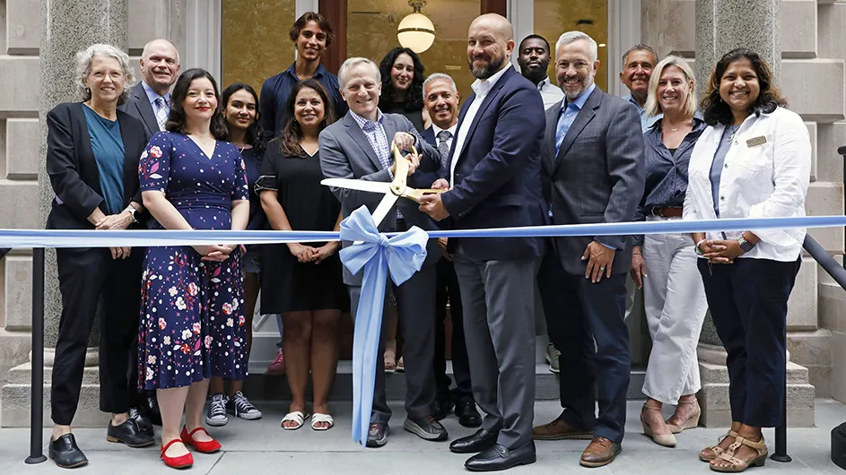 Fifteen university staff members and students smile and pose as Dean Josef Sorett and Executive VP David Greenberg hold oversized ceremonial scissors ready to cut a blue ribbon in front of a building entrance at Columbia University's first all-electric residence hall. 