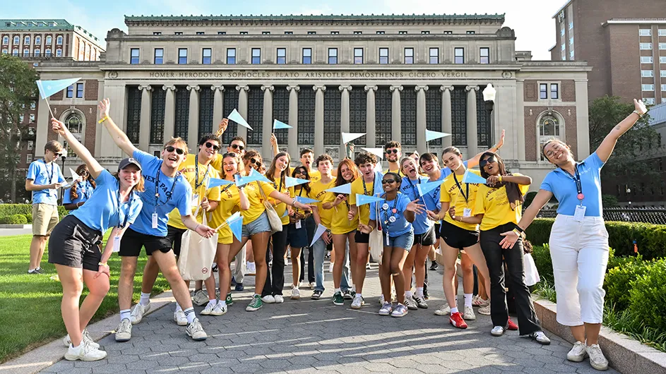 Several university students in light blue and yellow shirts while holding Columbia pennants pose and cheer with Butler Library in the background at Columbia University on a warm sunny day. 
