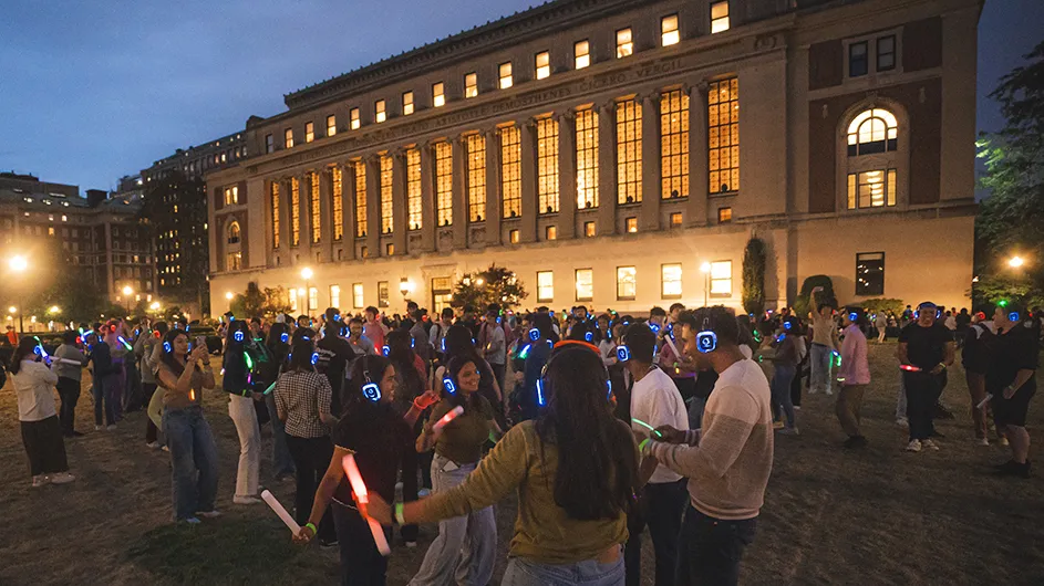 Dozens of university students wearing headphones smile and dance to music on South Lawn in front of Columbia University's illuminated Butler Library at night.