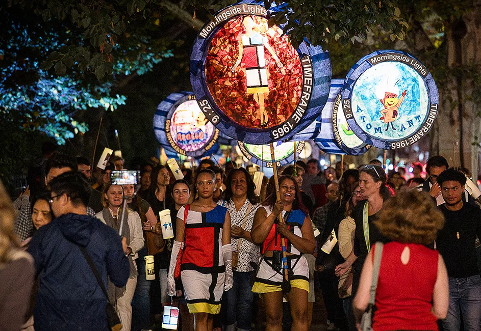Several people walking with two women, wearing Mondrian art color block dresses, on a parade carry colorful illuminated handmade lanterns on a warm night in New York City.