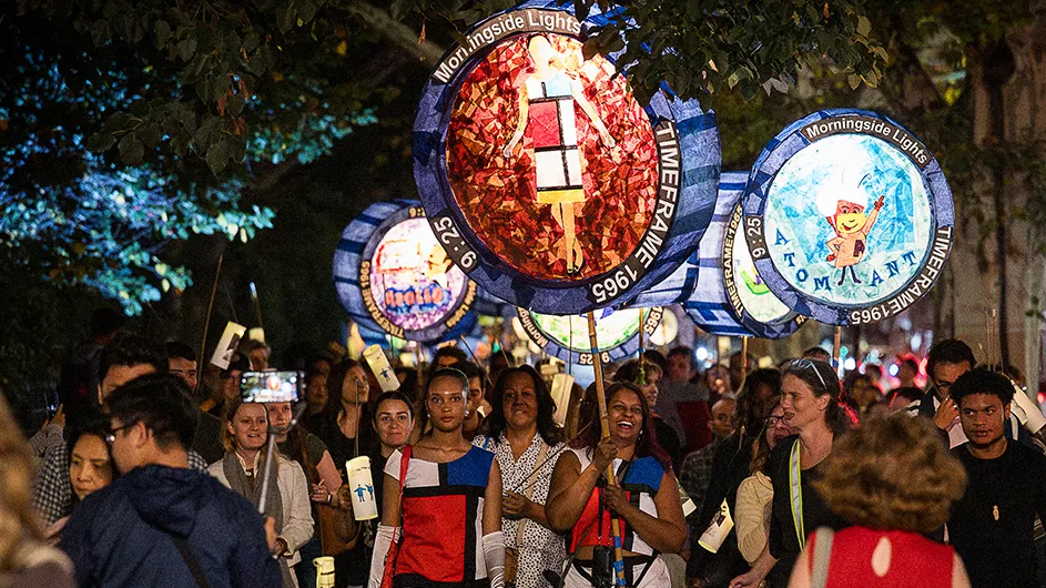 Several people walking with two women, wearing Mondrian art color block dresses, on a parade carry colorful illuminated handmade lanterns on a warm night in New York City.