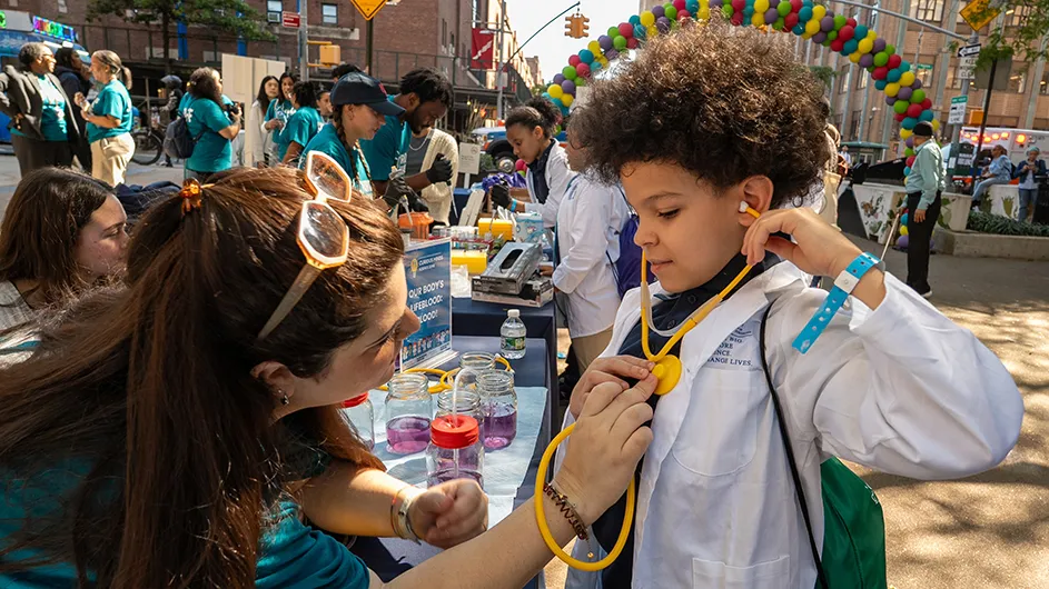 An elementary school boy in white lab coat listens to his heartbeat as a Columbia University employee holds a stethoscope to his heart at an outdoor event for school kids at Columbia University Irving Medical Center on a sunny day. 