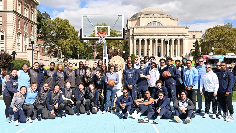 Several women's and men's basketball players dressed in Columbia uniforms smile and pose with Columbia's lion mascot and with Acting President Claire Shipman, who is smiling and holding a basketball, on South Lawn in front of a basketball backboard and Low Library in the background on a sunny day at Columbia University.  
