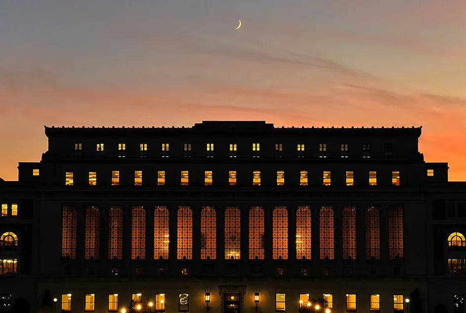 Columbia University's Butler Library illuminated at dusk with a crescent moon in the pink and gray sky above. 