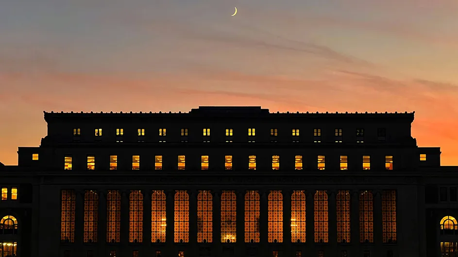 Columbia University's Butler Library illuminated at dusk with a crescent moon in the pink and gray sky above. 