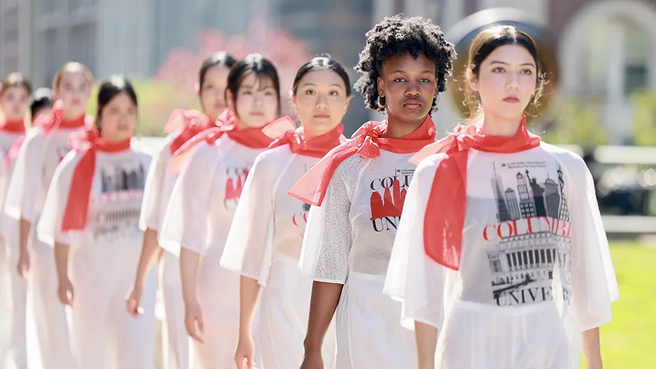 Several university students dressed in white and wearing red scarves walk in a Vietnamese heritage fashion runway show on Revson Plaza at Columbia University on a sunny day.