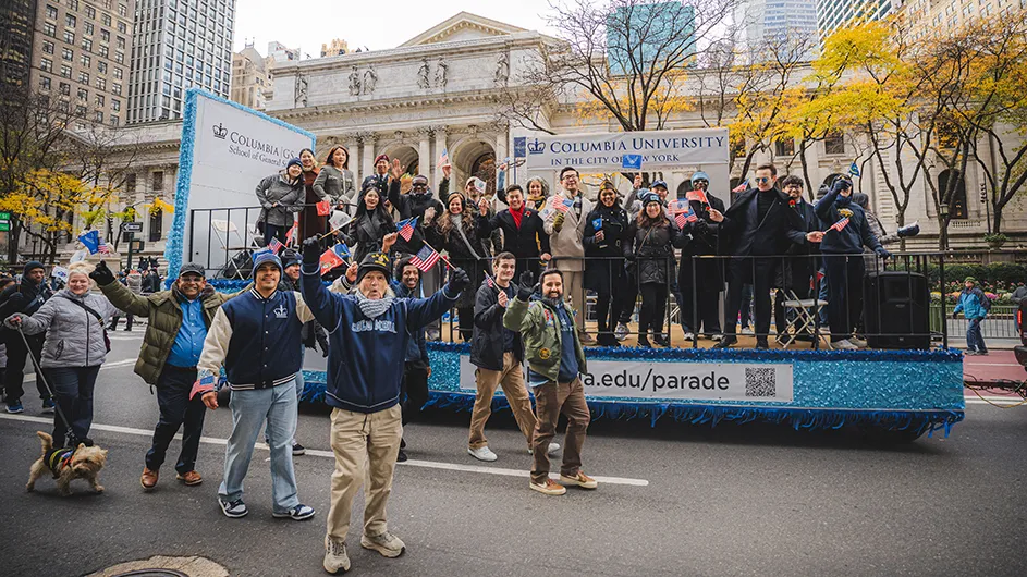Several Columbia University students, faculty, and alumni wave and smile aboard and alongside Columbia General Studies’ float in the New York City Veterans Day Parade, in front of the New York Public Library on a cloudy fall day. 
