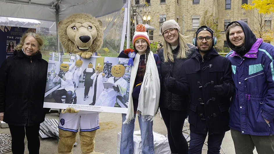 Five university students and faculty members smile and pose with Columbia's lion mascot holding up a photo of them with jack-o'-lanterns as heads at Columbia University Irving Medical Center.