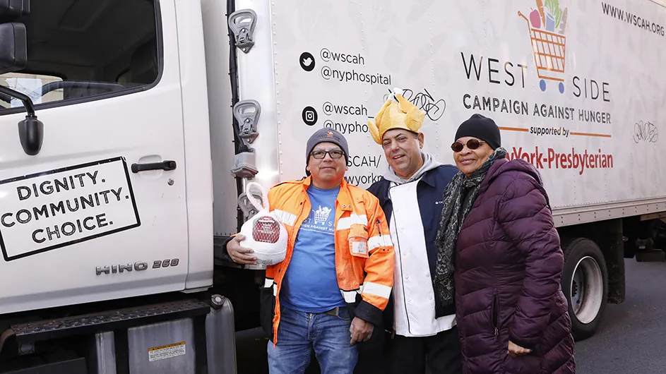 Two Columbia employees smile and pose with a West Side Campaign Against Hunger employee holding a frozen turkey next to a truck at a Thanksgiving turkey donation event on 114th Street at Columbia University.