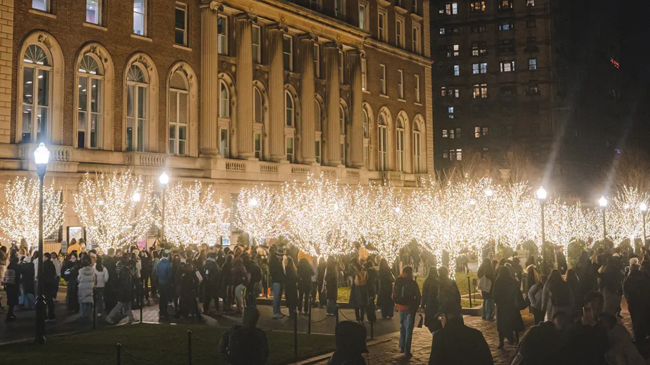 A crowd of university students gathers on lighted holiday tree-lined College Walk with Pulitzer Hall in the background at Columbia University at night.