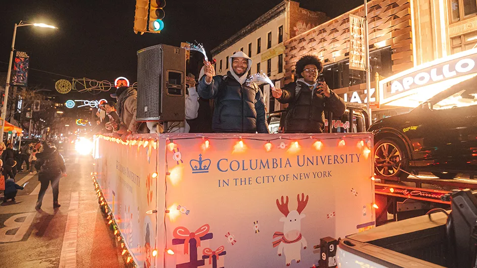 Several people in warm coats smile and cheer from the Columbia University float on 125th Street with spectators and the Apollo Theater in the background during the Harlem Holiday Lights parade.  