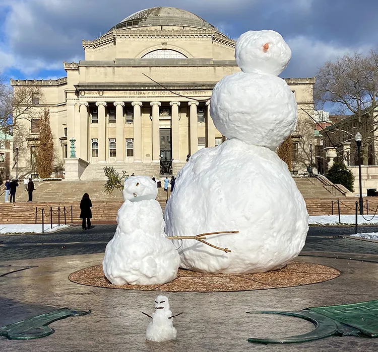 A snow family of three on the Sundial with Low Library in the background on a sunny day at Columbia University.