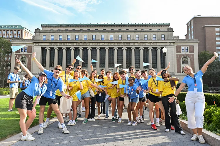 Several university students in light blue and yellow shirts while holding Columbia pennants pose and cheer with Butler Library in the background at Columbia University on a warm sunny day.