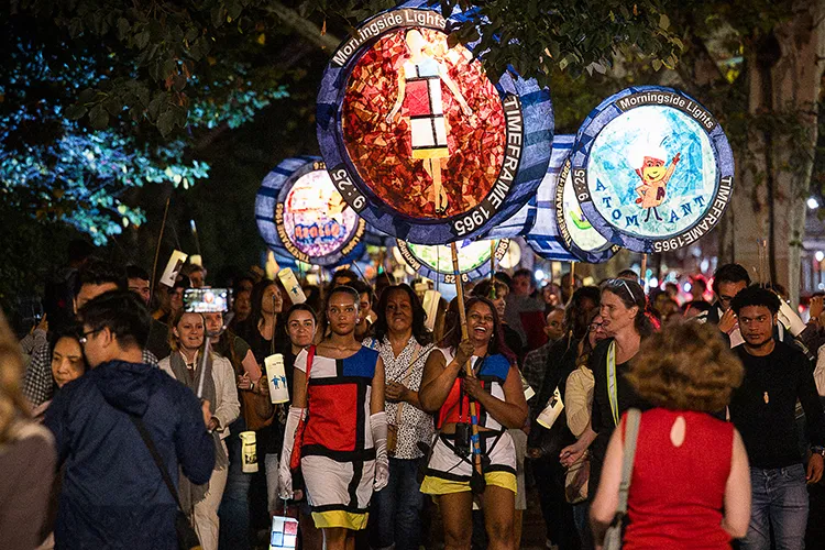 Several people walking with two women, wearing Mondrian art color block dresses, on a parade carry colorful illuminated handmade lanterns on a warm night in New York City.