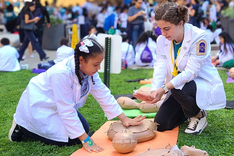 An elementary school girl in white lab coat performs chest compressions to a CPR manikin with a Columbia University employee at an outdoor event for school kids at Columbia University Irving Medical Center on a sunny day. 
