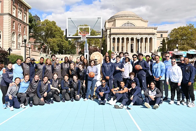Several women's and men's basketball players dressed in Columbia uniforms smile and pose with Columbia's lion mascot and with Acting President Claire Shipman, who is smiling and holding a basketball, on South Lawn in front of a basketball backboard and Low Library in the background on a sunny day at Columbia University.  