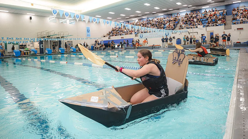 A university student wearing a life vest paddles a cardboard boat at Columbia University's Uris Pool with a large cheering crowd in the bleachers nearby. 