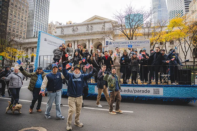 Several Columbia University students, faculty, and alumni wave and smile aboard and alongside Columbia General Studies’ float in the New York City Veterans Day Parade, in front of the New York Public Library on a cloudy fall day. 