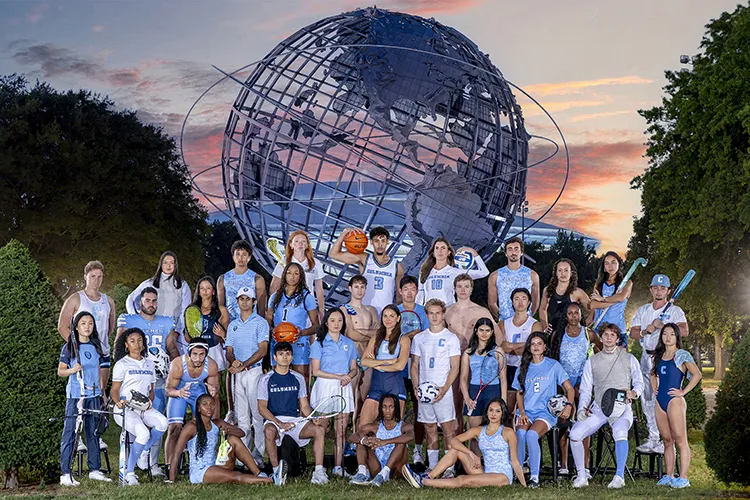 Thirty-one university student athletes in Columbia University sports uniforms pose in front of the Unisphere in Flushing Meadows Corona Park, Queens, at dusk. 