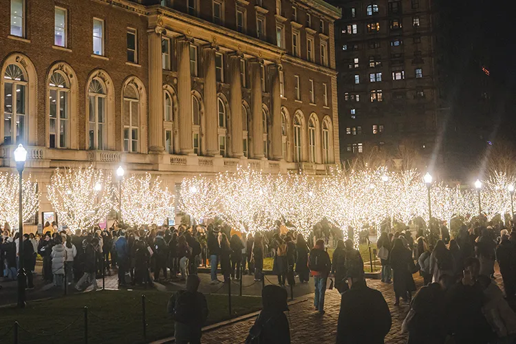 A crowd of university students gathers on sparkly-lighted holiday tree-lined College Walk with Pulitzer Hall in the background at Columbia University at night.