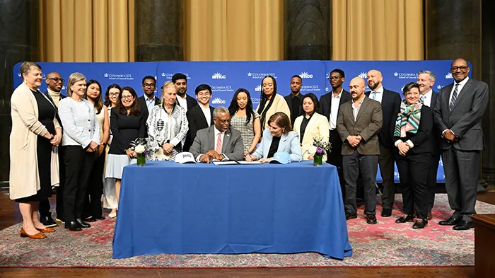 A group of students, faculty, and administrators, gather to watch an agreement being signed. 
