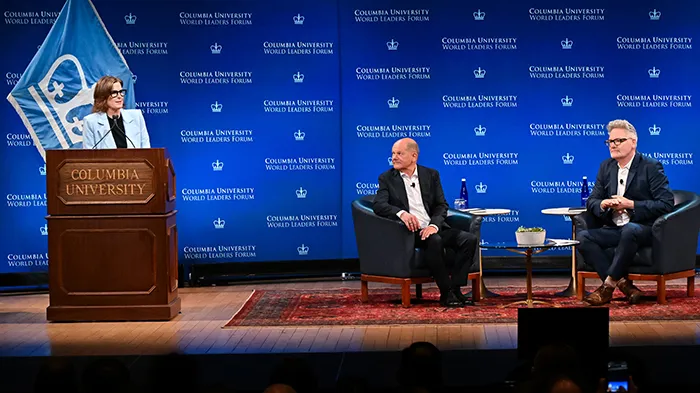 A woman at a podium alongside two men sitting down on a stage. 