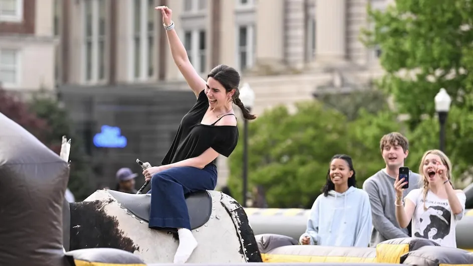Columbia University student riding a mechanical bull while other students look on. 