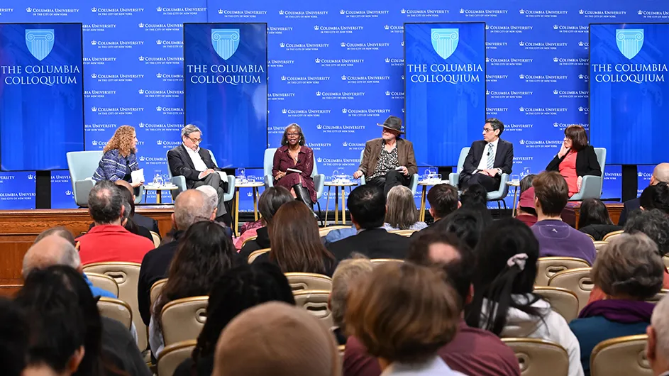 An audience watches a panel discussion with six people on a stage with “The Columbia Colloquium” blue and white banners in the background